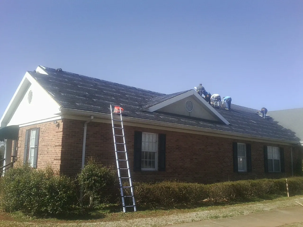 Crew tearing shingles off a brick ranch roof with a ladder set — Metal Roof Repair work by our crew in Severance