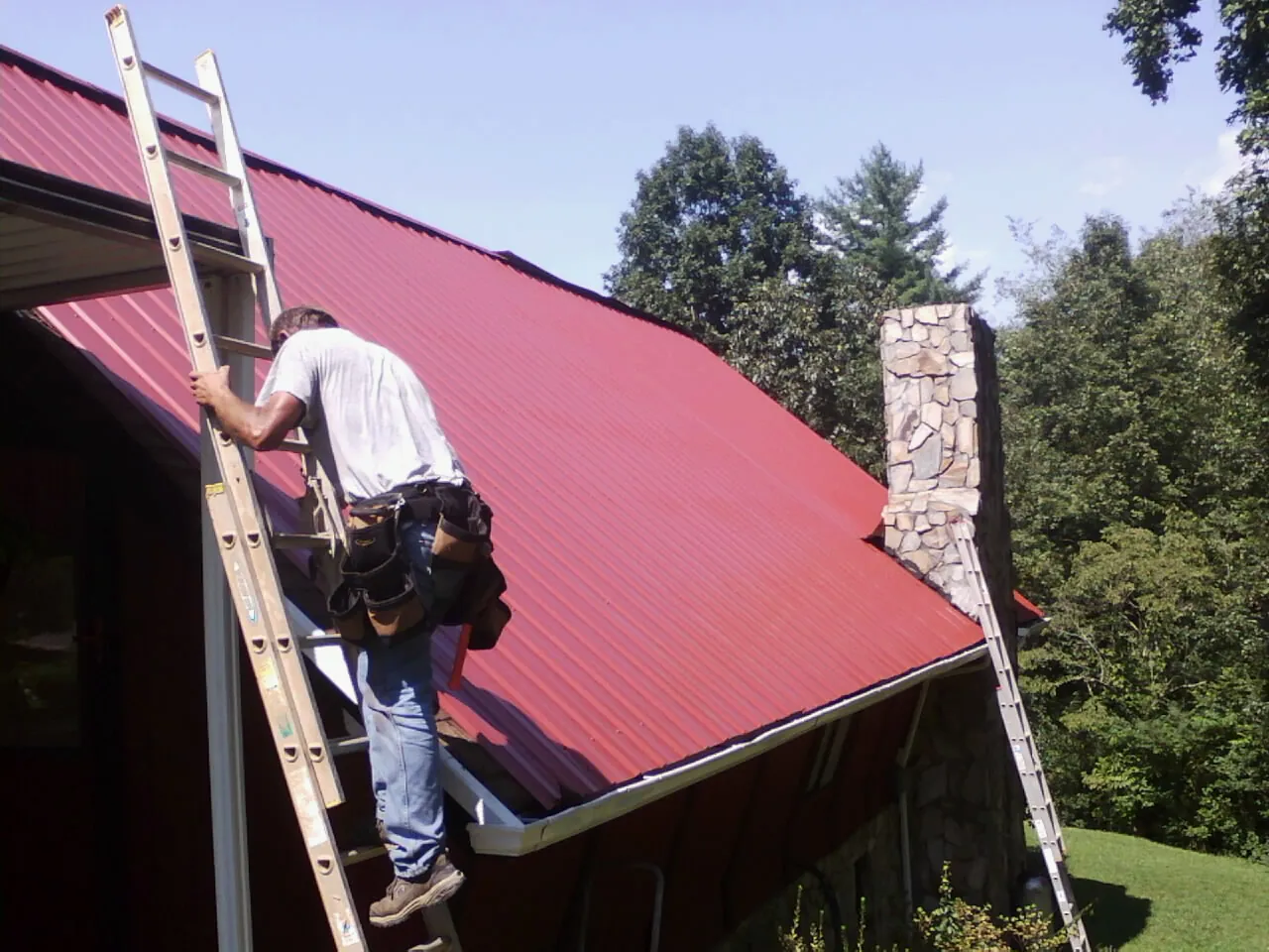 Roofer on a ladder beside a red metal roof and stone chimney — Soffit & Fascia Repair by Roofing Brothers in Severance